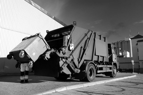 A black-and-white photograph shows a large commercial waste collection vehicle parked on a paved surface adjacent to a corrugated metal building. The truck has a hydraulic arm extending to lift a large, rectangular skip container filled with waste materials, which appears to be made of metal with a smooth, painted surface. The container is tilted slightly forward, ready for emptying. A worker wearing reflective safety trousers is standing beside the container, observing the process. The background includes industrial structures, possibly storage tanks or silos, with a cloudy sky overhead. The scene illustrates typical rubbish removal operations that a service like Commercial Waste Removal Brent Cross might undertake in the area, with the vehicle situated in a commercial or industrial setting, reflecting proximity to Brent Cross or nearby London boroughs.