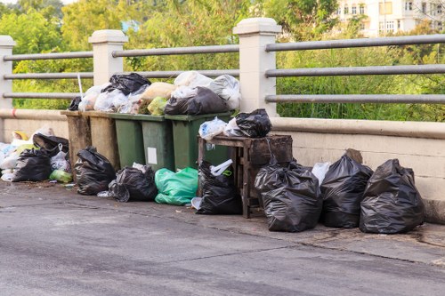 A collection of black, green, and white plastic rubbish bags, some partially filled and tied, are placed on the pavement and against a concrete barrier on a street. The barrier features evenly spaced metal posts and horizontal bars, which separate the walkway from a background of green trees and London-style buildings. Nearby, there are also some cardboard boxes and a few loose items among the bags, indicating waste collected for removal in the Brent Cross area. The scene is illuminated by natural daylight, with no visible people present. This image depicts typical outdoor rubbish disposal and highlights the need for professional waste removal services such as those provided by Commercial Waste Removal Brent Cross, ensuring proper collection and environmentally responsible disposal of waste materials in urban environments.
