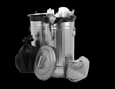 A large, round metal rubbish bin with a fitted lid sits on a flat surface, filled to the brim with crumpled paper and plastic waste, with some contents overflowing and draping over the top edge. A black plastic garbage bag leans against the bin on the left side, tied at the top, and a smaller white paper bag is positioned to the right, all set against a plain dark background. The scene suggests waste collection or disposal in a professional waste removal context, typical of commercial waste management services in areas such as Brent Cross, north London. The metallic surface of the bin appears shiny with subtle reflections, highlighting its sturdy construction, while the waste materials display a mix of textures, from crumpled paper to soft plastic. This setup captures the typical state of waste awaiting removal, emphasizing a neat and organized approach to rubbish disposal for businesses or properties in the local area.