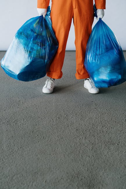 A person dressed in orange overalls and white sneakers is standing on a gray paved surface, holding two large blue plastic rubbish bags filled with waste. The bags are semi-transparent, revealing vari