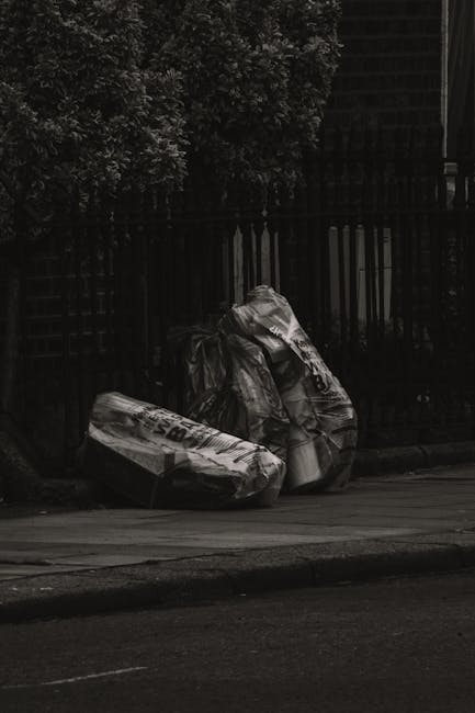 A person dressed in orange overalls and white sneakers is standing on a gray paved surface, holding two large blue plastic rubbish bags filled with waste. The bags are semi-transparent, revealing vari