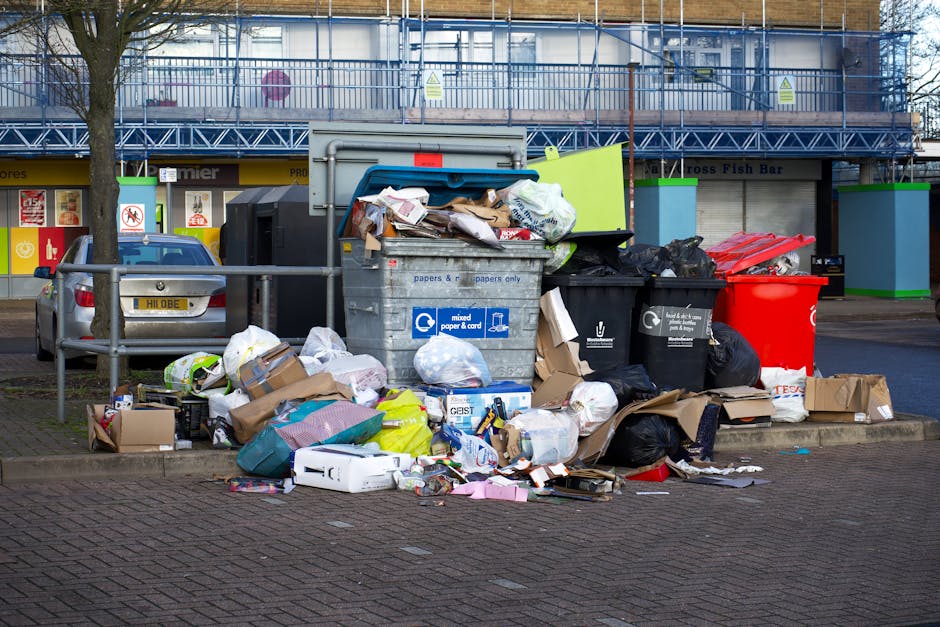 A public street scene featuring a collection of discarded rubbish situated around a concrete block and a metal waste container with a rounded, dark finish. The rubbish includes various cardboard boxes