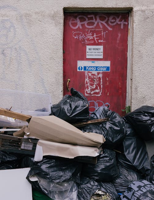 A collection of black rubbish bags and discarded cardboard and wooden packaging materials are piled in front of a red metal door set into a concrete wall, which is marked with graffiti tags in white a