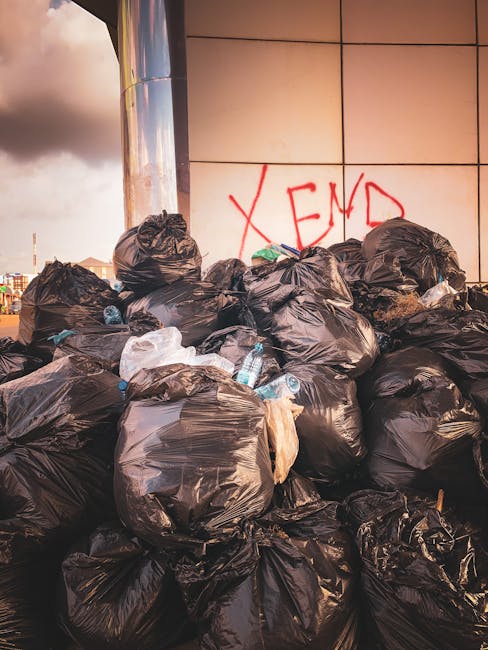 A collection of black rubbish bags and discarded cardboard and wooden packaging materials are piled in front of a red metal door set into a concrete wall, which is marked with graffiti tags in white a