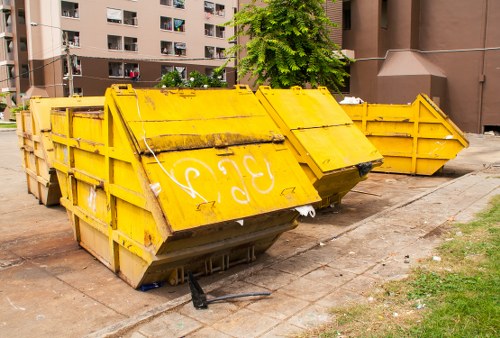 A row of large industrial skip containers painted bright yellow with reinforced metal sides and hinged lids, positioned on a paved outdoor area adjacent to a residential apartment building in an urban setting. The skips are partially filled with waste, and some lids are securely closed while others are slightly ajar or tilted open, revealing dark debris and mixed rubbish inside. The containers are aligned side by side, with the closest one tilted forward, displaying a sloped top and a sturdy black wheel supporting its angled front. The surrounding environment includes a patch of grass near the pavement, and the background features multi-storey residential blocks with balconies and windows, typical of a busy town setting. These skips are situated along a concrete pathway, possibly part of a commercial waste removal operation by Commercial Waste Removal Brent Cross, serving waste collection needs for local businesses across the area, including postcode district NW4 in Brent Cross. The scene is lit with natural daylight, indicating a clear weather day, emphasizing the functional, utilitarian nature of the waste management equipment.