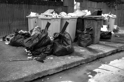 A row of four large wheeled rubbish bins, with three in a light beige colour and one in dark grey, positioned on a paved driveway or sidewalk near a wooden fence. The bins are overflowing with various waste items, including crumpled papers and cardboard, with some waste spilling over their edges. Surrounding the bins are numerous black plastic rubbish bags, some tied and others open, containing various discarded materials. The bags are stacked and scattered on the ground, with a few leaning against the bins. The environment appears to be an outdoor domestic or commercial area, with a weathered concrete surface and a wooden fence in the background. The image relates to waste collection and rubbish removal services, exemplifying typical waste accumulation requiring professional waste management, as offered by Commercial Waste Removal Brent Cross, servicing local businesses and residents in the Brent Cross area.