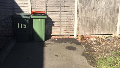 The image shows two wheeled rubbish bins placed on a concrete driveway in front of a wooden fence; one bin is dark green with a closed lid, and the other is light green with an orange lid, both marked with numbers; the driveway is shaded on one side and exposed to sunlight on the other, with visible dark and light patches of stain on the surface. The surrounding environment includes a gravelly area with some small plants near the fence, which appears weathered and slightly uneven, typical of a residential or small commercial setting in Brent Cross, where Commercial Waste Removal Brent Cross offers rubbish removal services. The scene captures a typical rubbish collection point, emphasizing the importance of proper waste management for local properties.