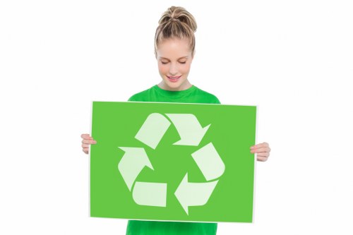 A young woman with blonde hair tied in a ponytail, wearing a bright green long-sleeved shirt, is smiling and holding a large rectangular green sign featuring a white recycling symbol composed of three arrows forming a triangle. The background of the image is plain white, emphasizing her and the sign. This visual promotes recycling awareness, aligning with sustainable waste management practices often associated with rubbish removal services. The woman's cheerful expression and the bold green color of her shirt and sign convey a positive message about environmental responsibility. The recycling symbol on the sign is prominent and centrally positioned, making the message clear. This image could be relevant for a waste management company's campaign emphasizing eco-friendly disposal methods, particularly in areas like Brent Cross where local waste regulations encourage recycling and responsible rubbish removal. The clean, professional appearance of the image supports the values of proper waste sorting and environmental consciousness central to commercial waste removal services in the UK.